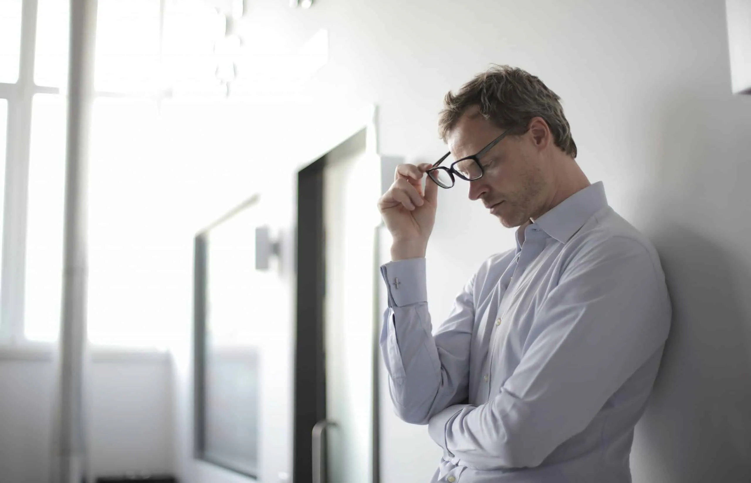 A stressed man leaning against a wall whilst taking his glasses off.